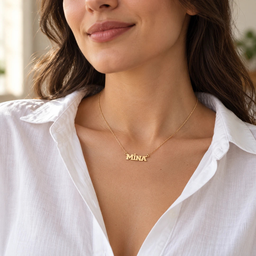 CHOICE Marcella Blok Yazılı İsim Kolye, MİNA kişiselleştirme örneği; 925 ayar gümüş üzerine 14 ayar altın kaplama; beyaz gömlekli kadın modelde minimal duruş